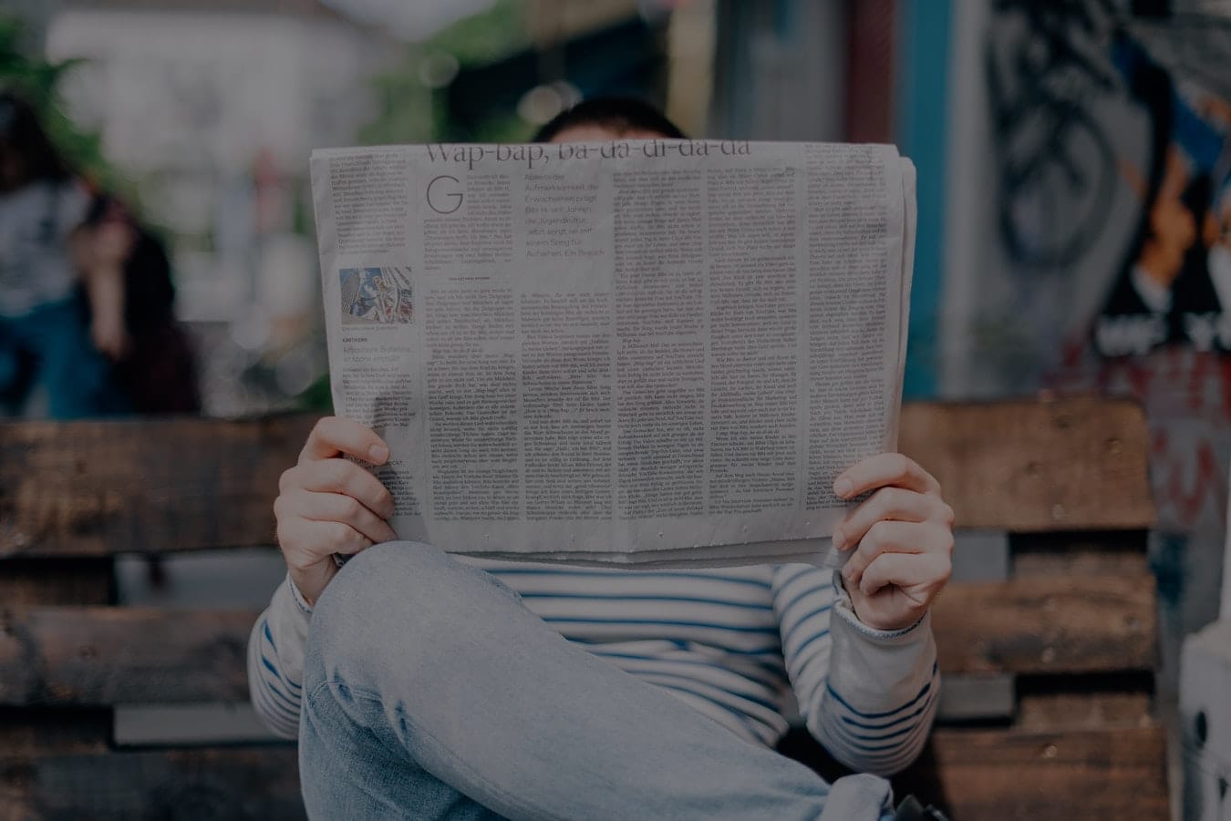 Man reading a newspaper seated at a bench.