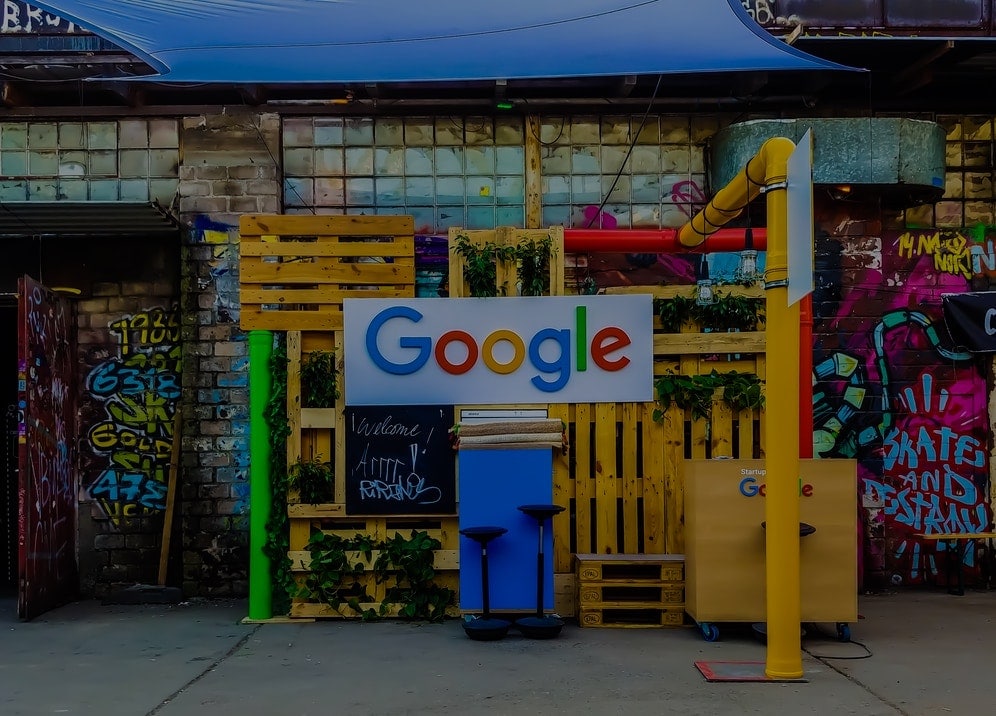 Street sidewalk view of the Google logo on a banner in front of arranged pallet boxes and graffiti.